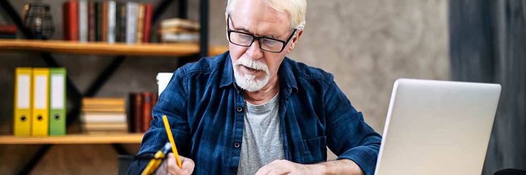 A senior man takes notes during an online class.