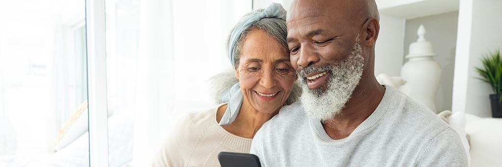A senior couple sits on a couch and looks at a phone together.