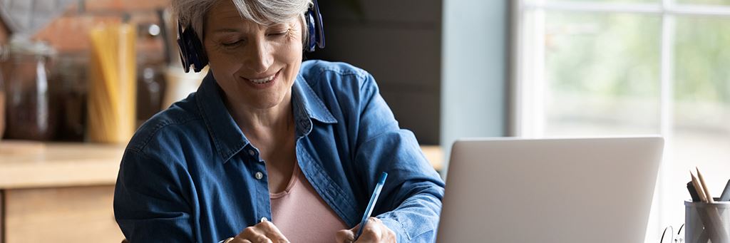 A woman wearing headphones takes notes while looking at her computer.