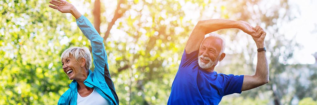 An older couple does a side stretch outside in a park.