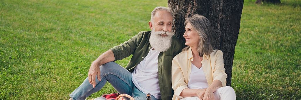 An older couple with gray hair leans on a tree in a park.