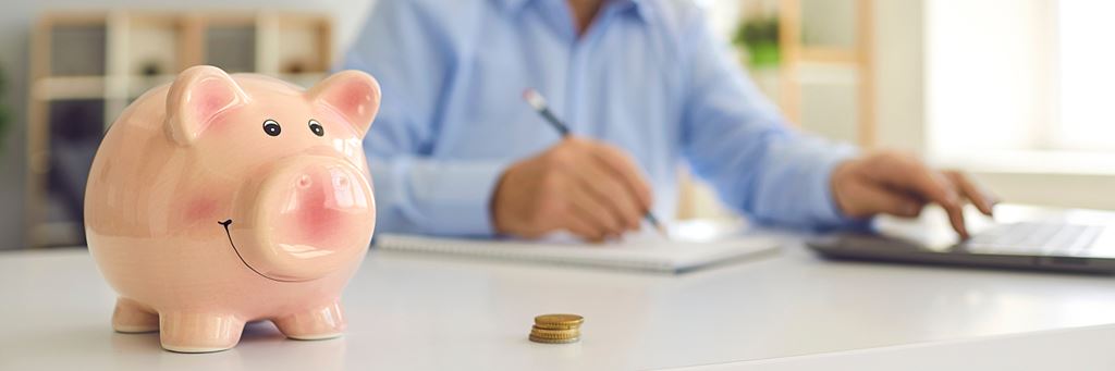 A piggy bank in the foreground next to a stack of coins with a person using a calculator.