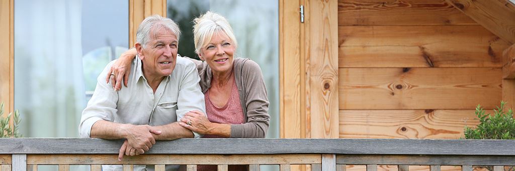 A senior couple stands on the front porch of their home.