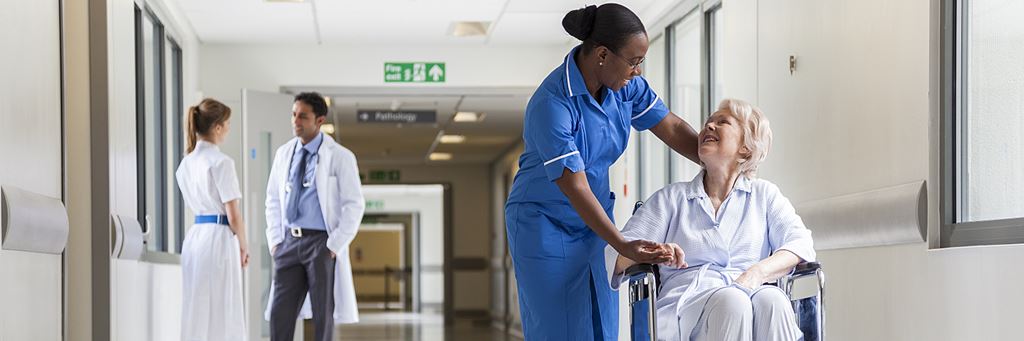 A nurse attends to a senior patient in a wheelchair in a hospital hallway.