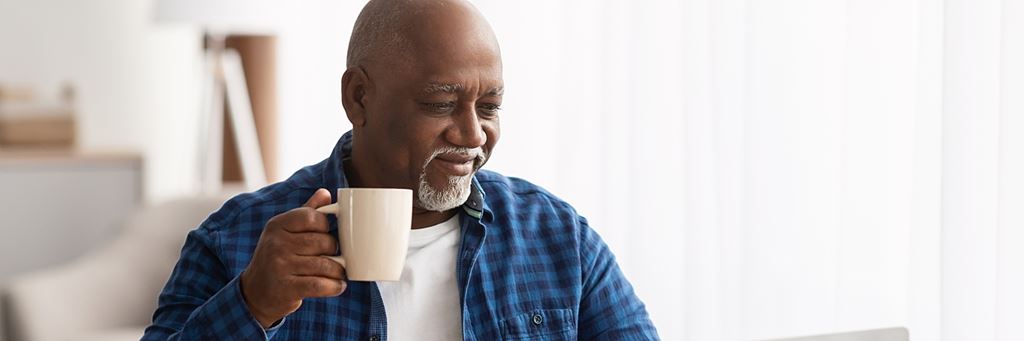An older man drinks a cup of coffee and works on a laptop.