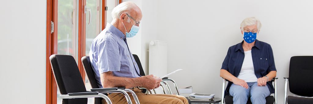 A senior man and woman wearing face masks wait in a doctor's office waiting room.