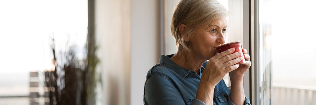 A pensive woman stands against a wall while looking out the window and sipping from a mug.