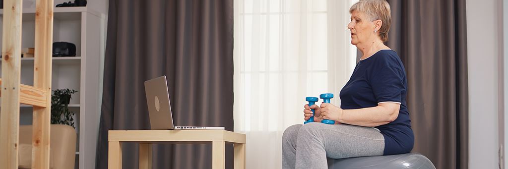 A senior woman exercises at home using an exercise ball and weights.