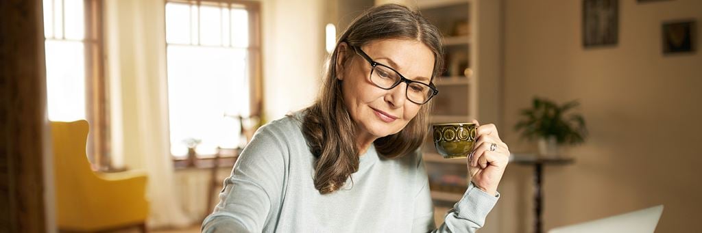 A senior woman holds a coffee cup and reviews paperwork.