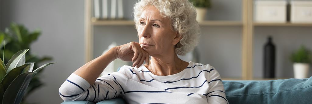 A thoughtful senior woman looks out the window while sitting on her living room couch.