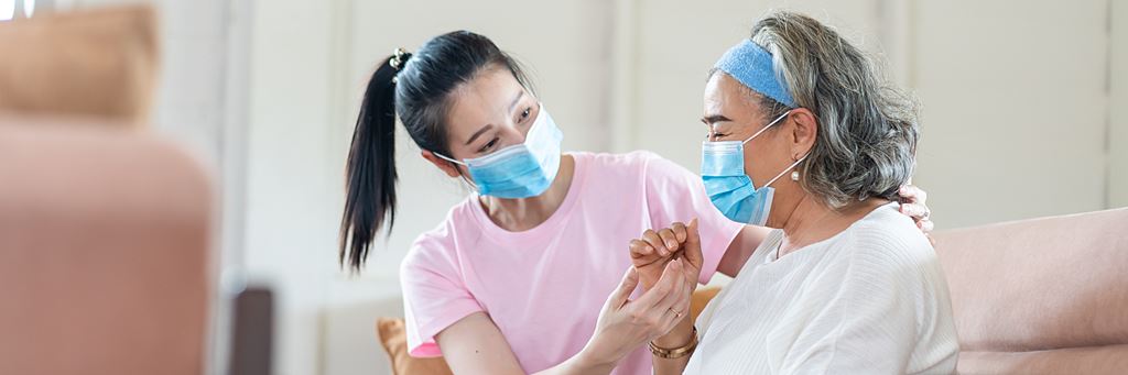 An in-home caregiver assists an elderly patient while they both wear face masks.